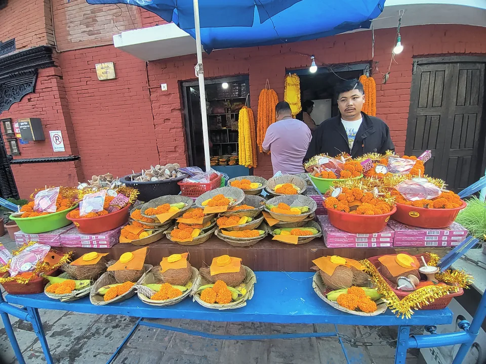 A stall at Kumbheshwar displays bowls of offerings and orange marigold garlands against a brick wall.